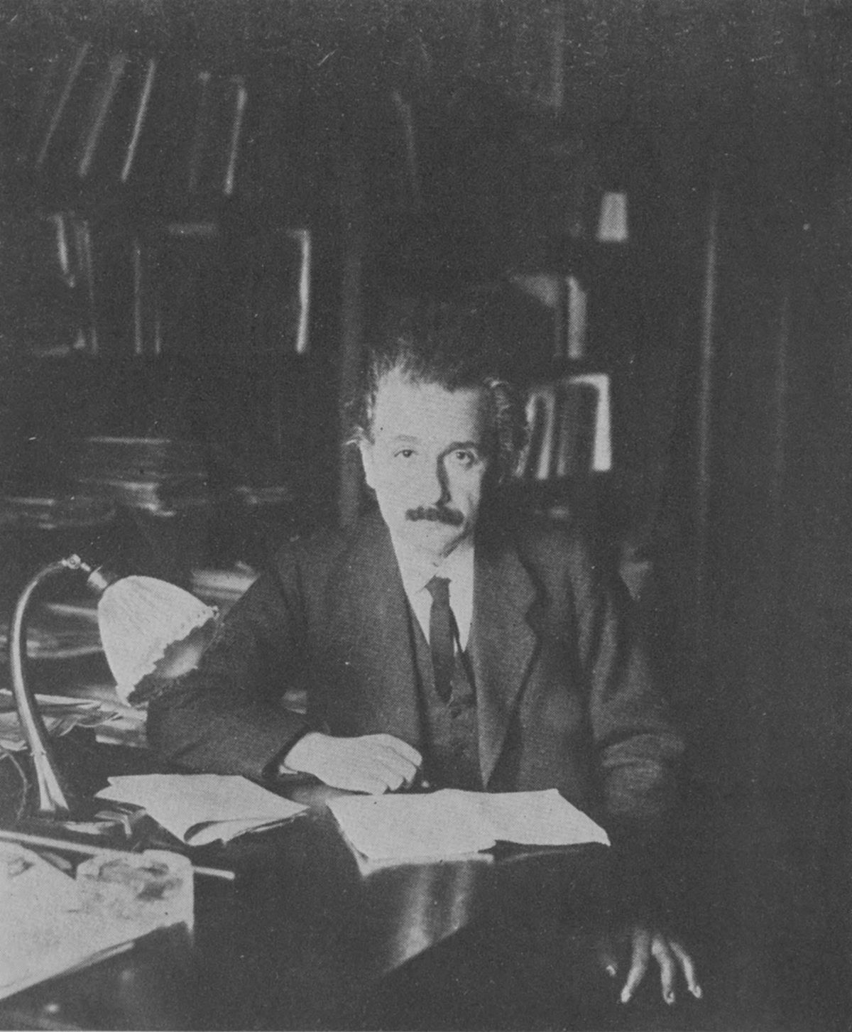 Black-and-white photo of Einstein with wild hair and mustache seated at a desk with papers and a lamp.